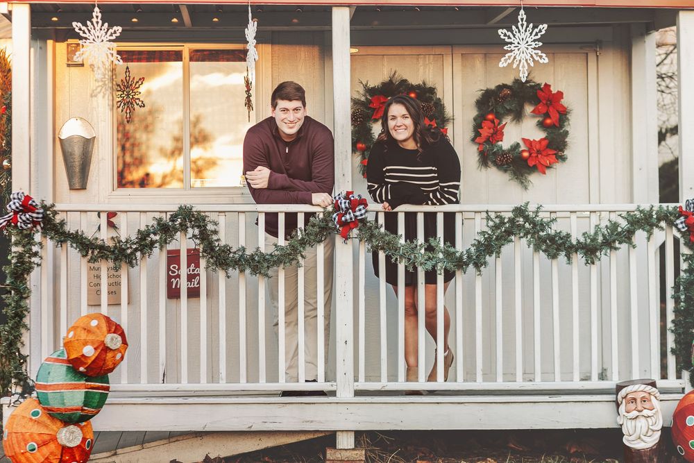 Couple smiling on a festively decorated porch with Christmas wreaths, garland, and holiday figurines.