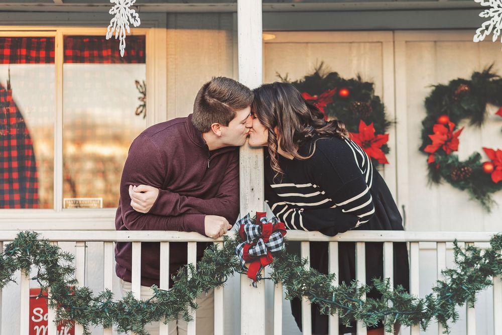 Couple sharing a kiss on a Christmas-decorated porch with wreaths and garland holiday decor.