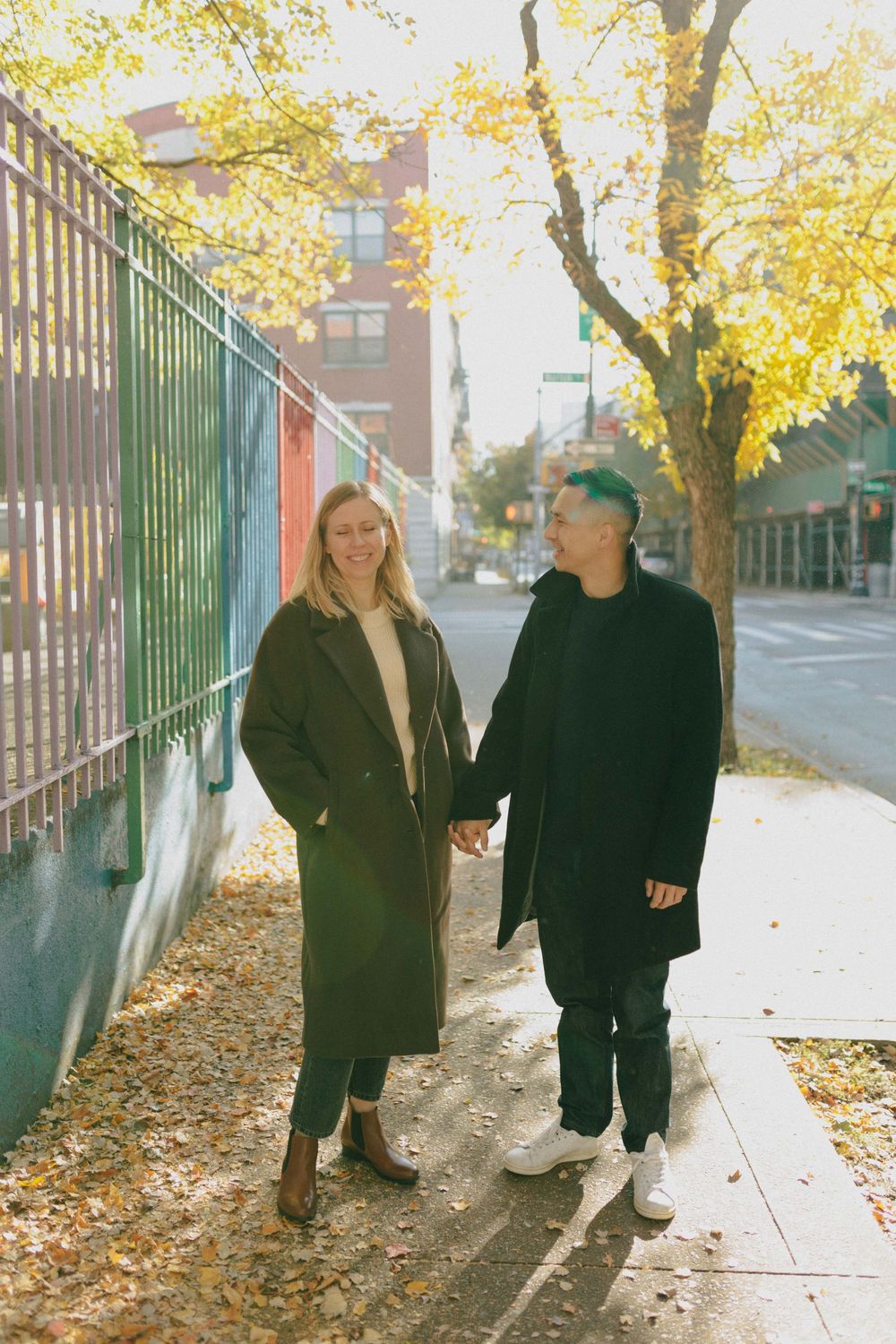 Couple holding hands on a sunny autumn sidewalk lined with golden leaves in the city.