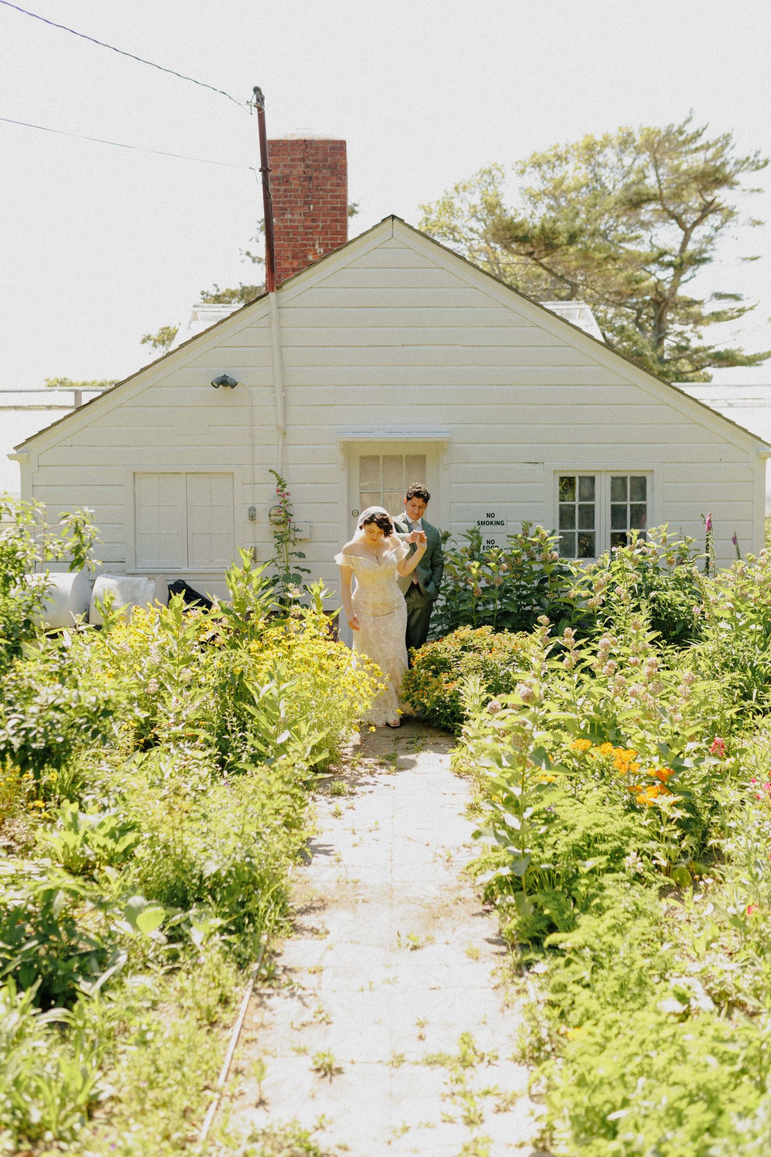 Bride and groom stand on a garden path surrounded by lush greenery in front of a white cottage home.
