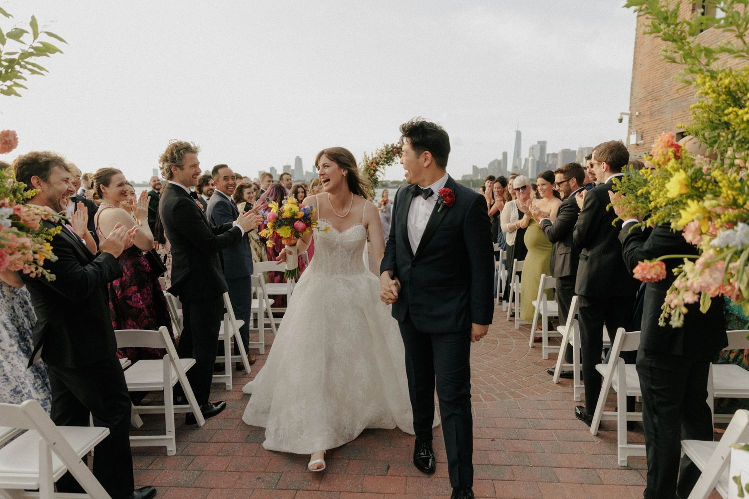 Bride and groom walk down outdoor wedding aisle, guests cheering, city skyline backdrop, colorful floral arrangements.
