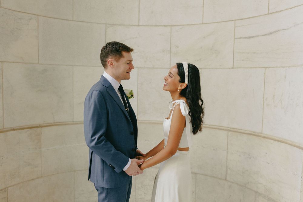 Bride and groom holding hands and smiling at each other against a curved stone wall.