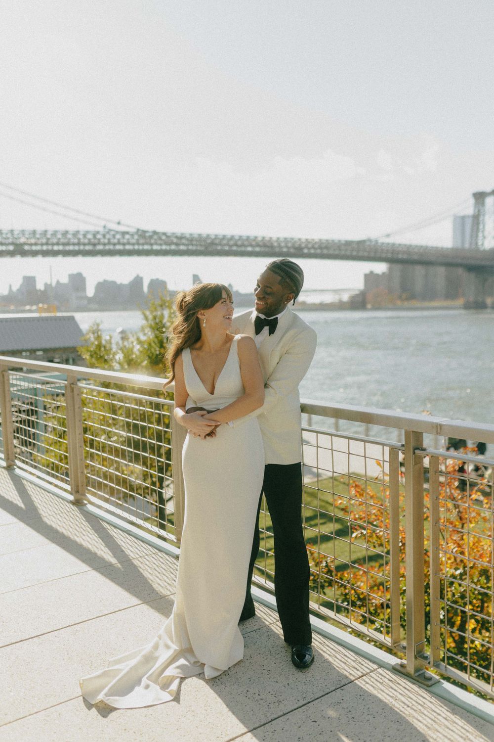 Bride and groom embracing on a waterfront terrace with the Brooklyn Bridge in the background.