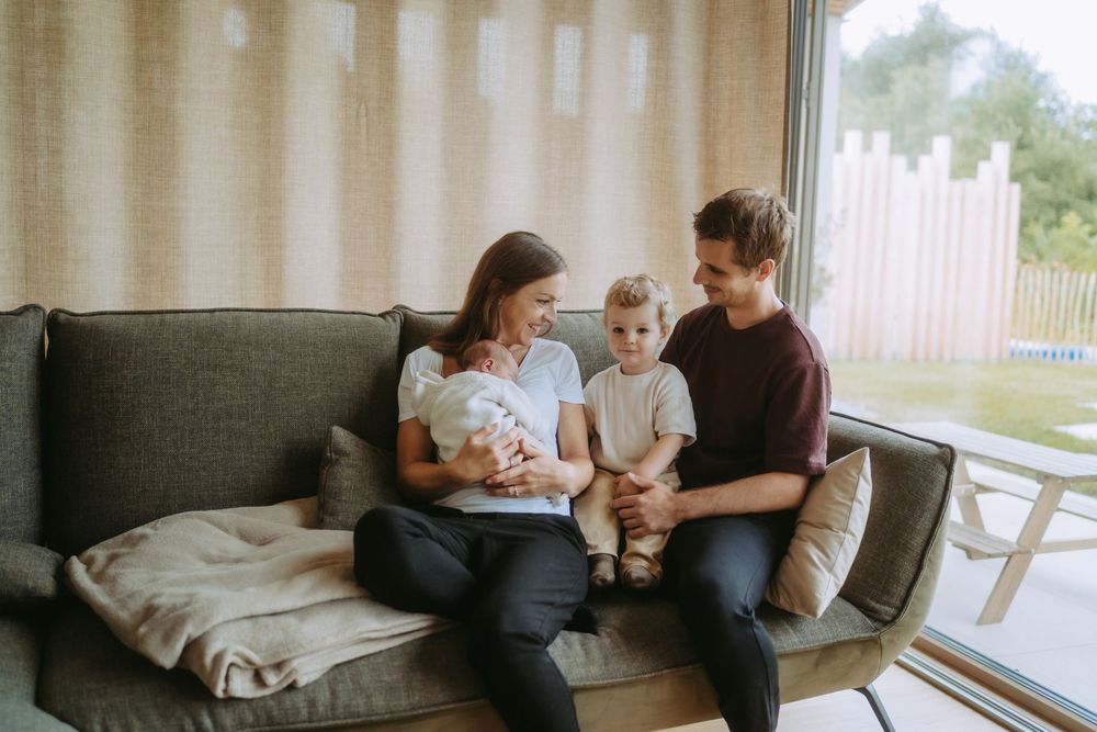Young family with newborn baby and toddler sitting together on a cozy gray sofa near a large window.