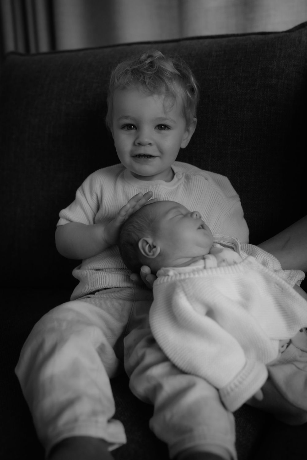 Toddler boy smiling while gently holding a sleeping newborn baby sibling in black and white photo.