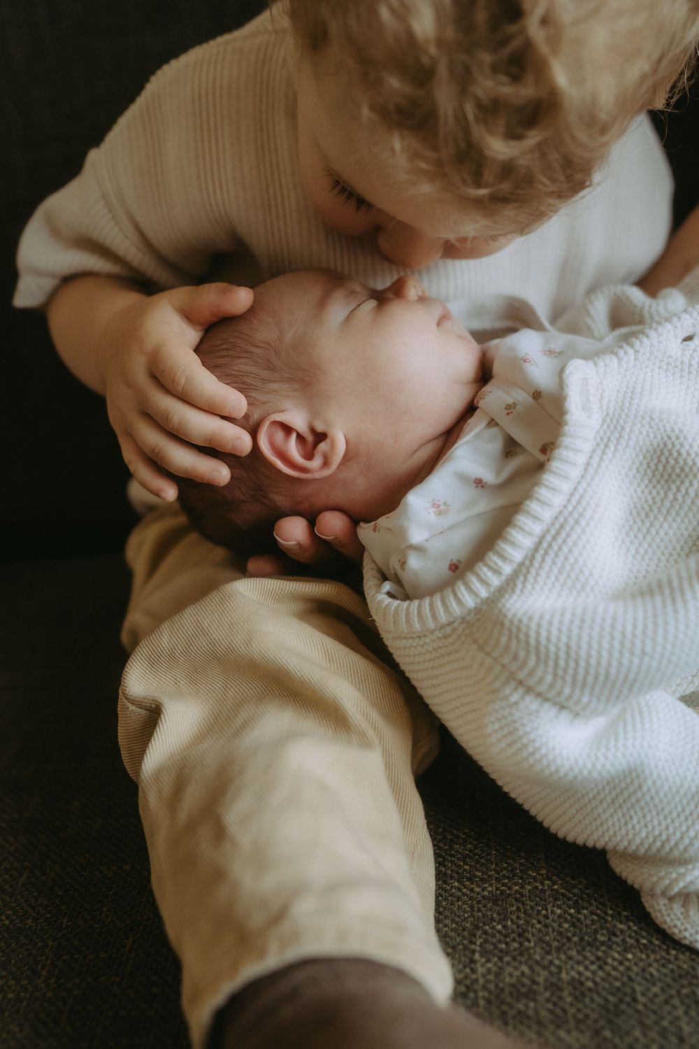Toddler gently kissing newborn baby sibling on the forehead while cradling their head with care.