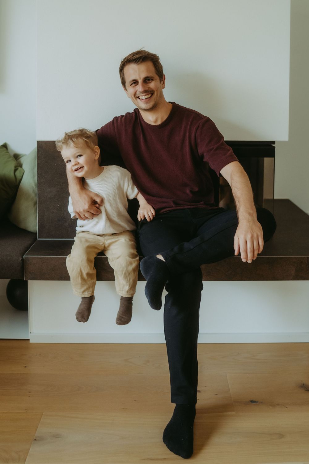 Smiling father sitting with toddler son on bench in modern living room, sharing a joyful moment together.