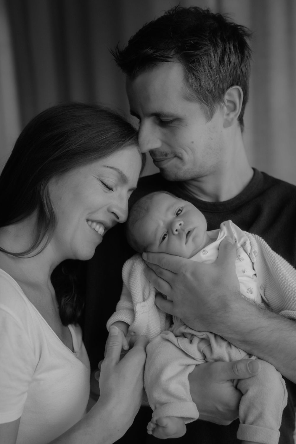 Black and white photo of happy parents smiling and cuddling their newborn baby together.