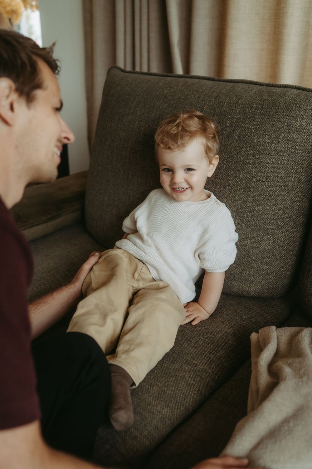 A smiling toddler in a white sweater and tan pants sits on a gray sofa while an adult man interacts with them.