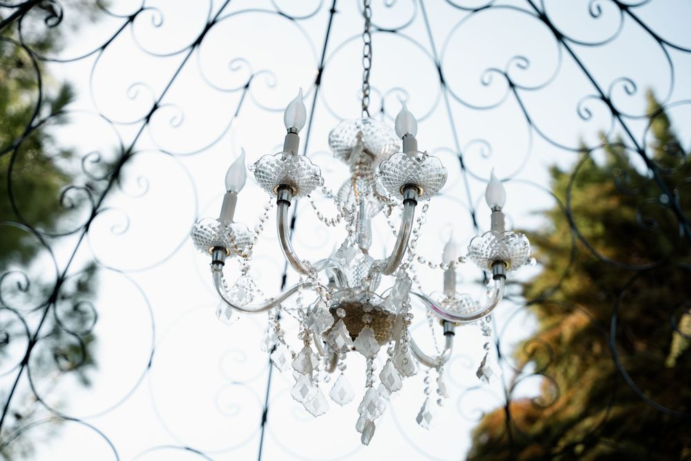 Elegant white crystal chandelier with candle arms hanging outdoors, framed by ornate black ironwork and lush green trees.