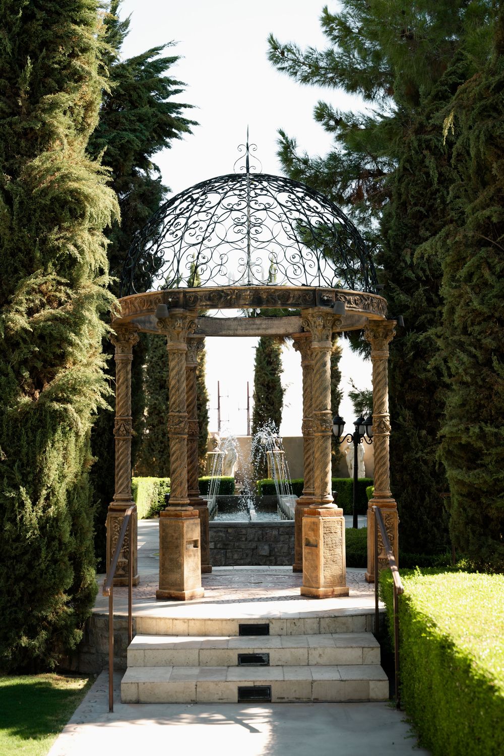 Ornate stone gazebo with wrought iron dome surrounded by cypress trees and manicured hedges in a formal garden.