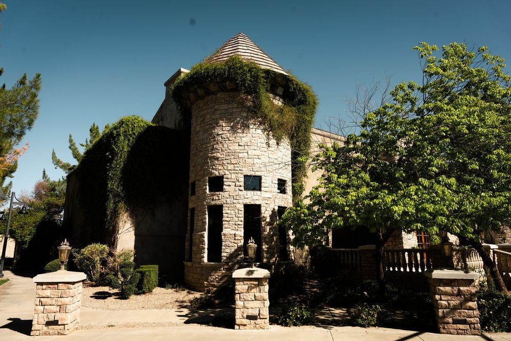 Stone castle-style building with turret, ivy-covered walls, and lush green trees under a clear blue sky.