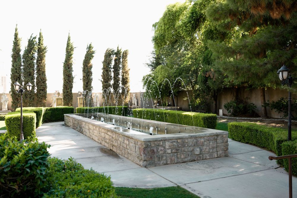 Elegant garden fountain with stone basin, manicured hedges, tall cypress trees, and lush greenery in a serene outdoor setting.