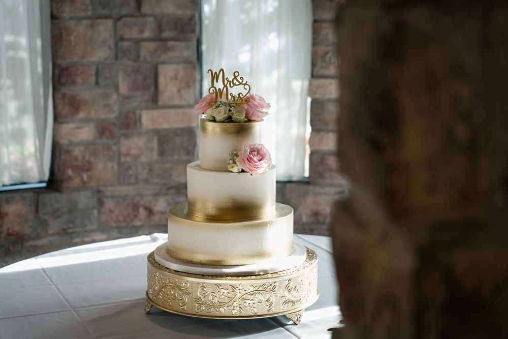 Elegant four-tier white and gold wedding cake with pink roses and Mr. & Mrs. topper on ornate gold stand.