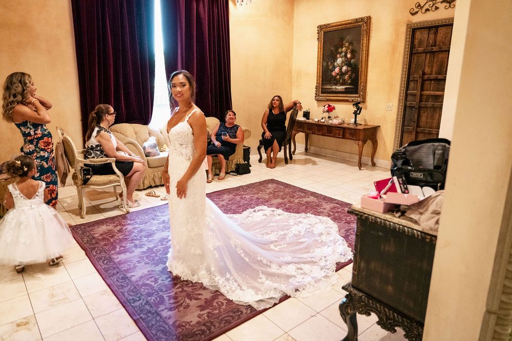 Bride in elegant lace wedding gown with long train stands in ornate room while guests admire her bridal look.