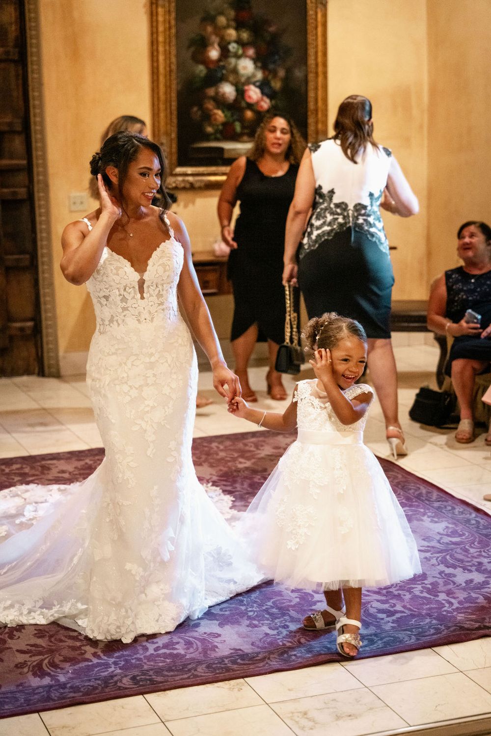 Bride in lace gown holds hands with flower girl in white dress, walking together at elegant wedding venue.