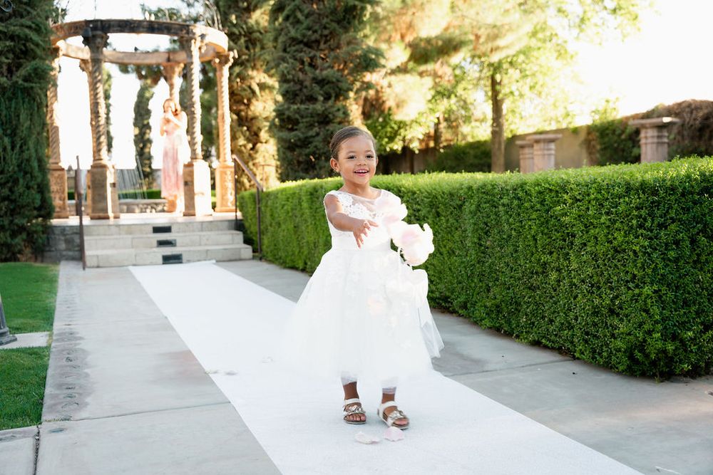Adorable flower girl in white dress walking down wedding aisle holding bouquet at outdoor garden ceremony.