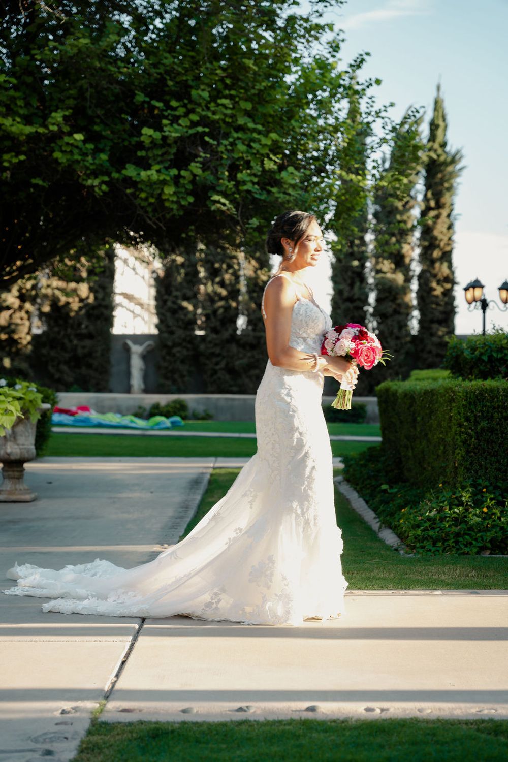 Elegant bride in white lace wedding gown holding pink bouquet standing in lush garden pathway.