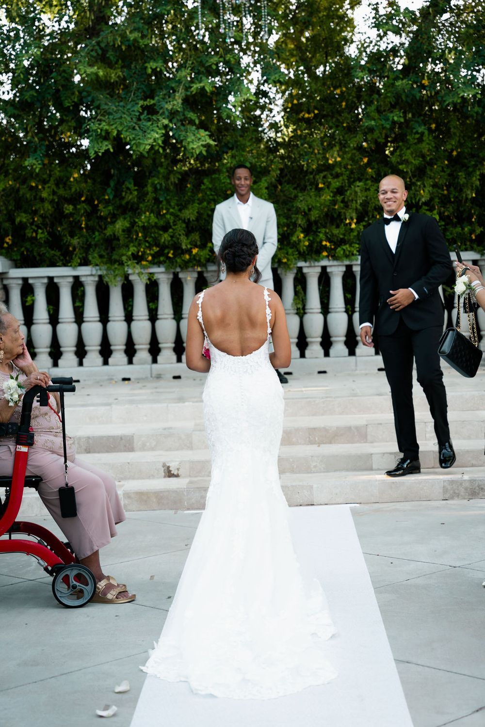 Bride in white backless gown walks down aisle toward groom in tuxedo at outdoor wedding ceremony.
