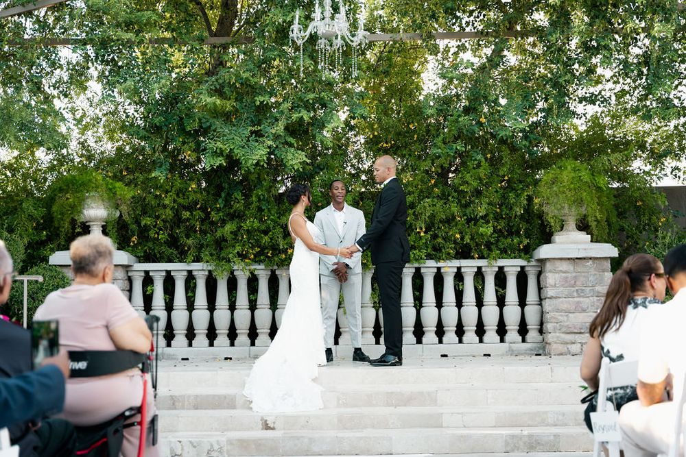 Outdoor wedding ceremony on stone steps with bride and groom at altar, lush green trees and white balustrade backdrop.
