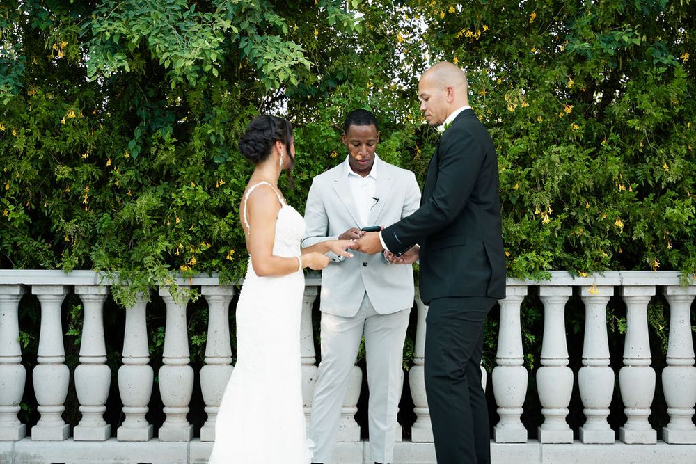 Outdoor wedding ceremony with bride, groom, and officiant exchanging vows on an elegant stone balcony surrounded by lush greenery.