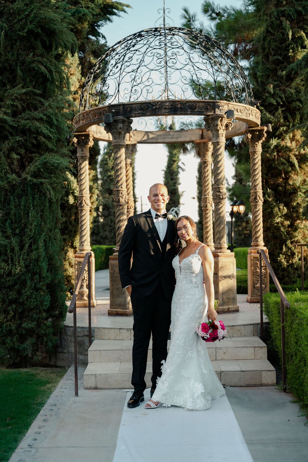 Bride and groom posing in front of ornate gazebo surrounded by lush greenery on their wedding day.