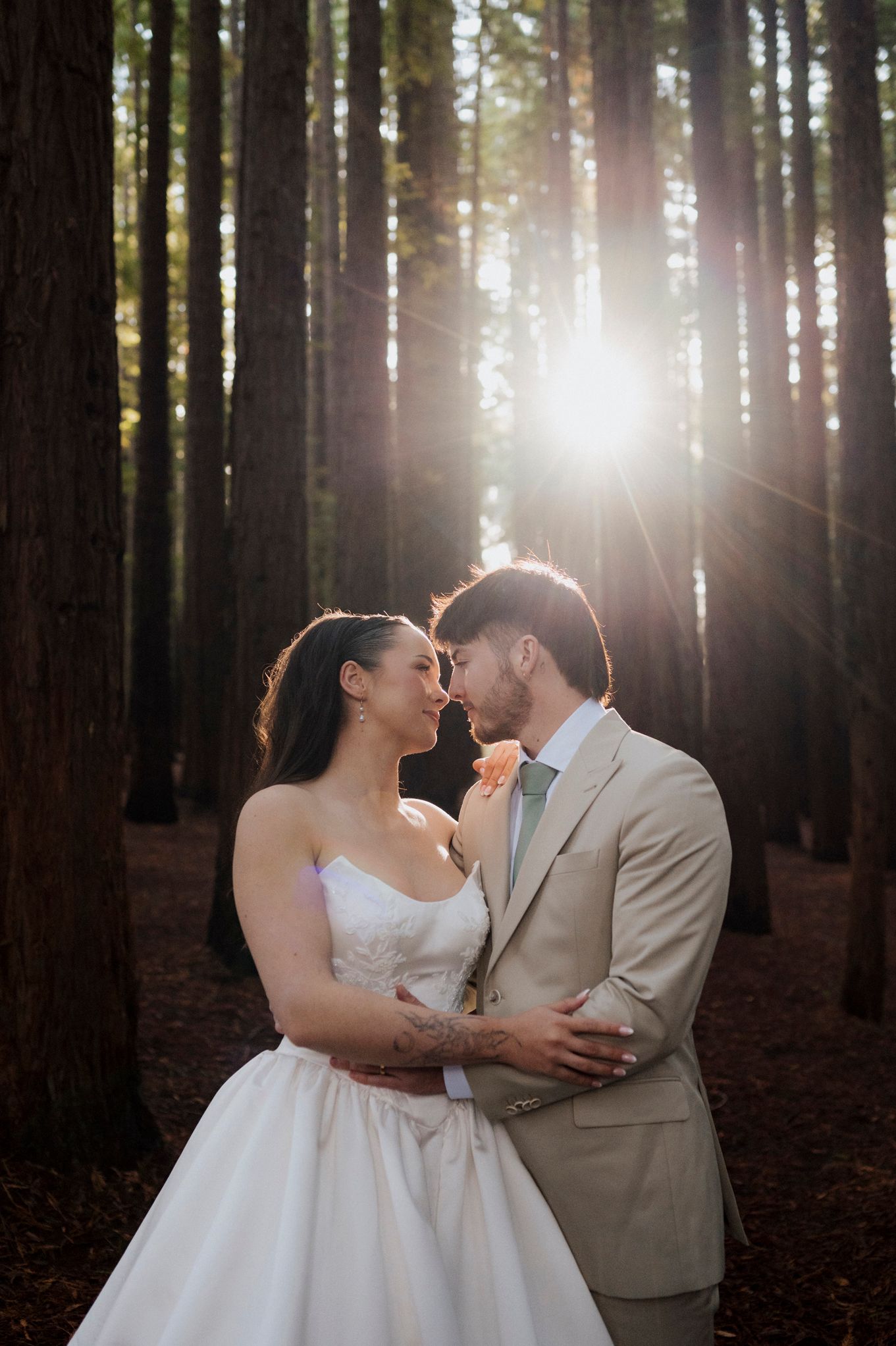 Bride and groom share a romantic moment in a sunlit redwood forest, with golden rays streaming through tall trees.