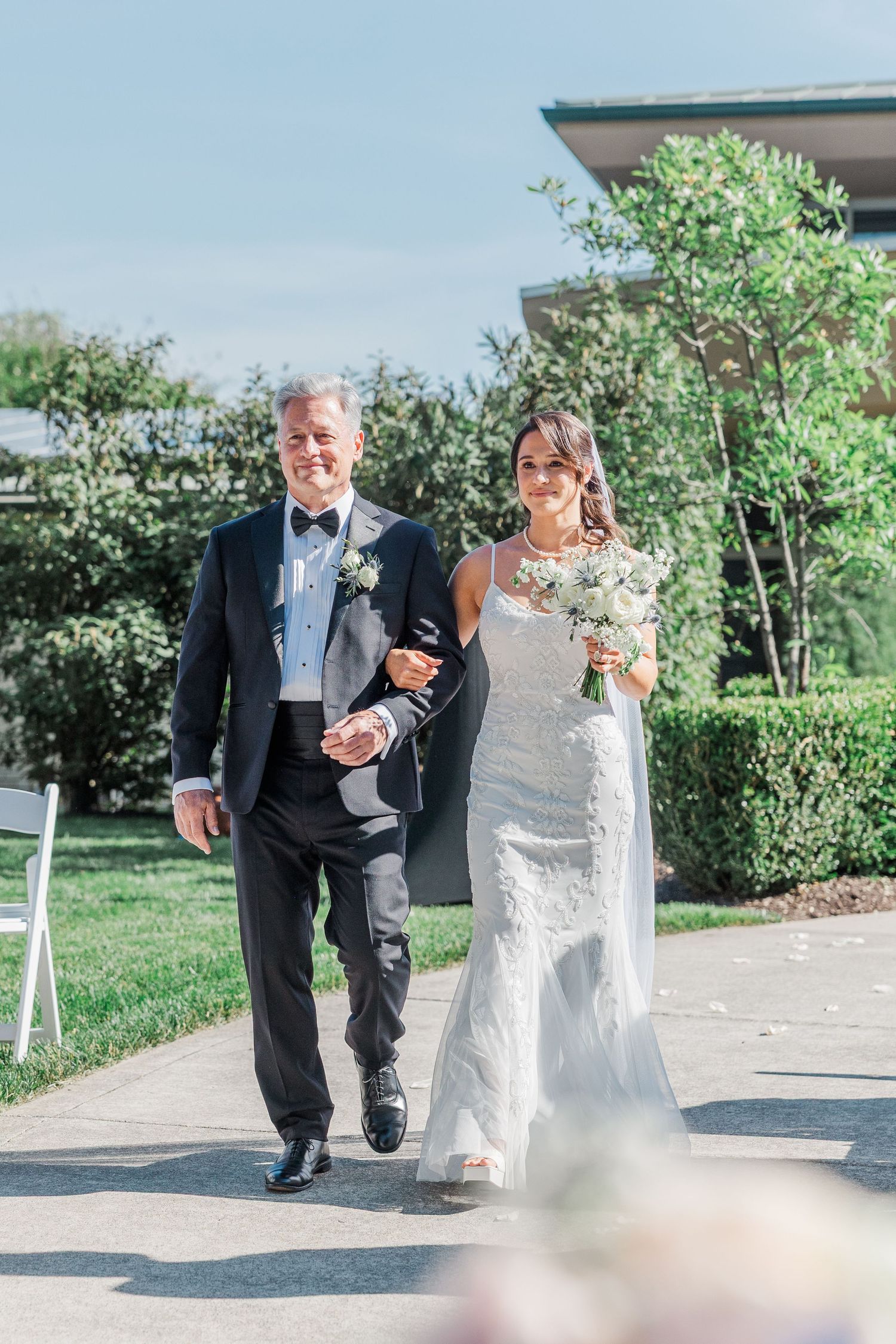 Bride in lace gown walks down outdoor aisle with father in tuxedo, holding white bouquet on sunny wedding day.