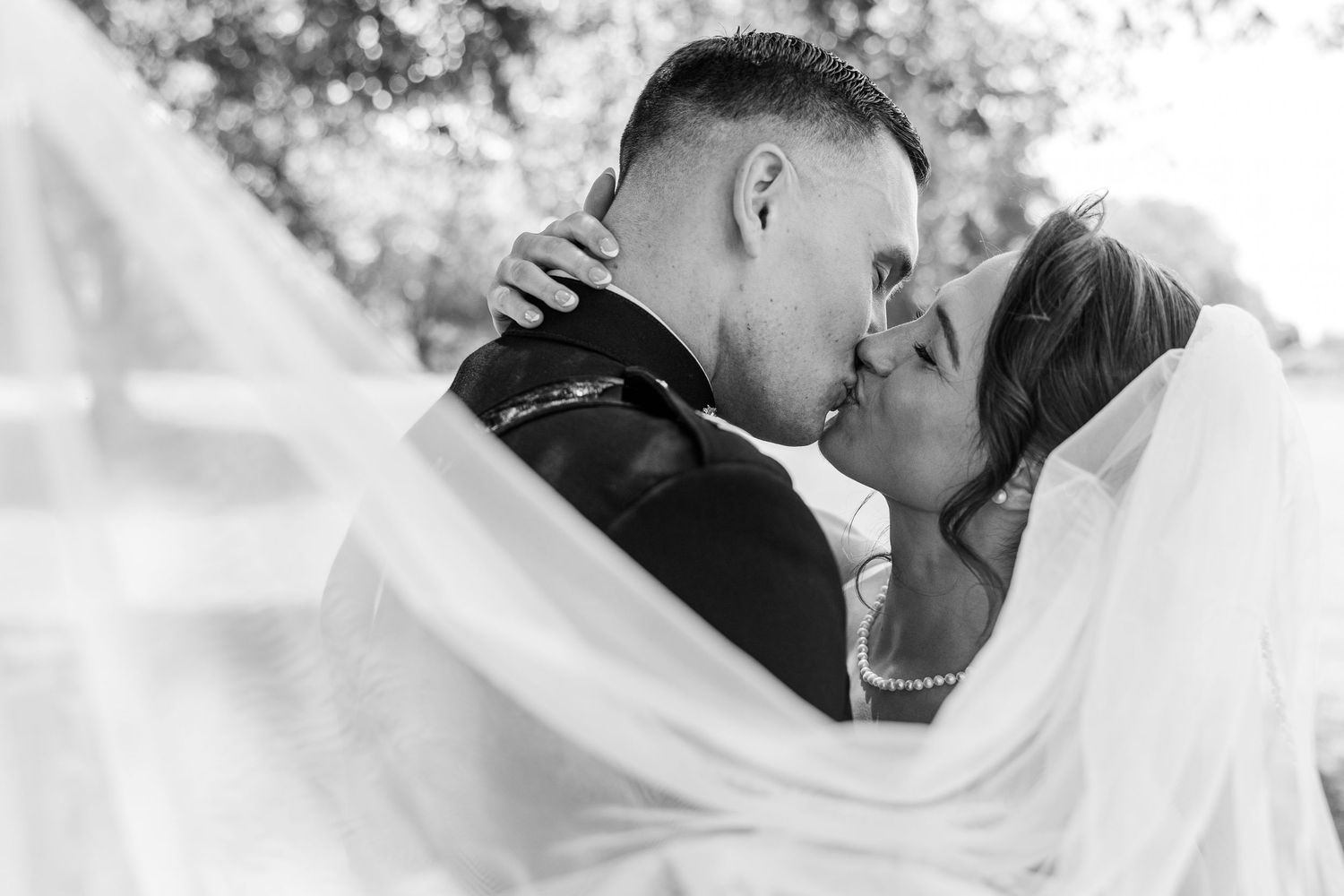 Black and white photo of a bride and groom sharing a romantic kiss under a flowing veil outdoors.