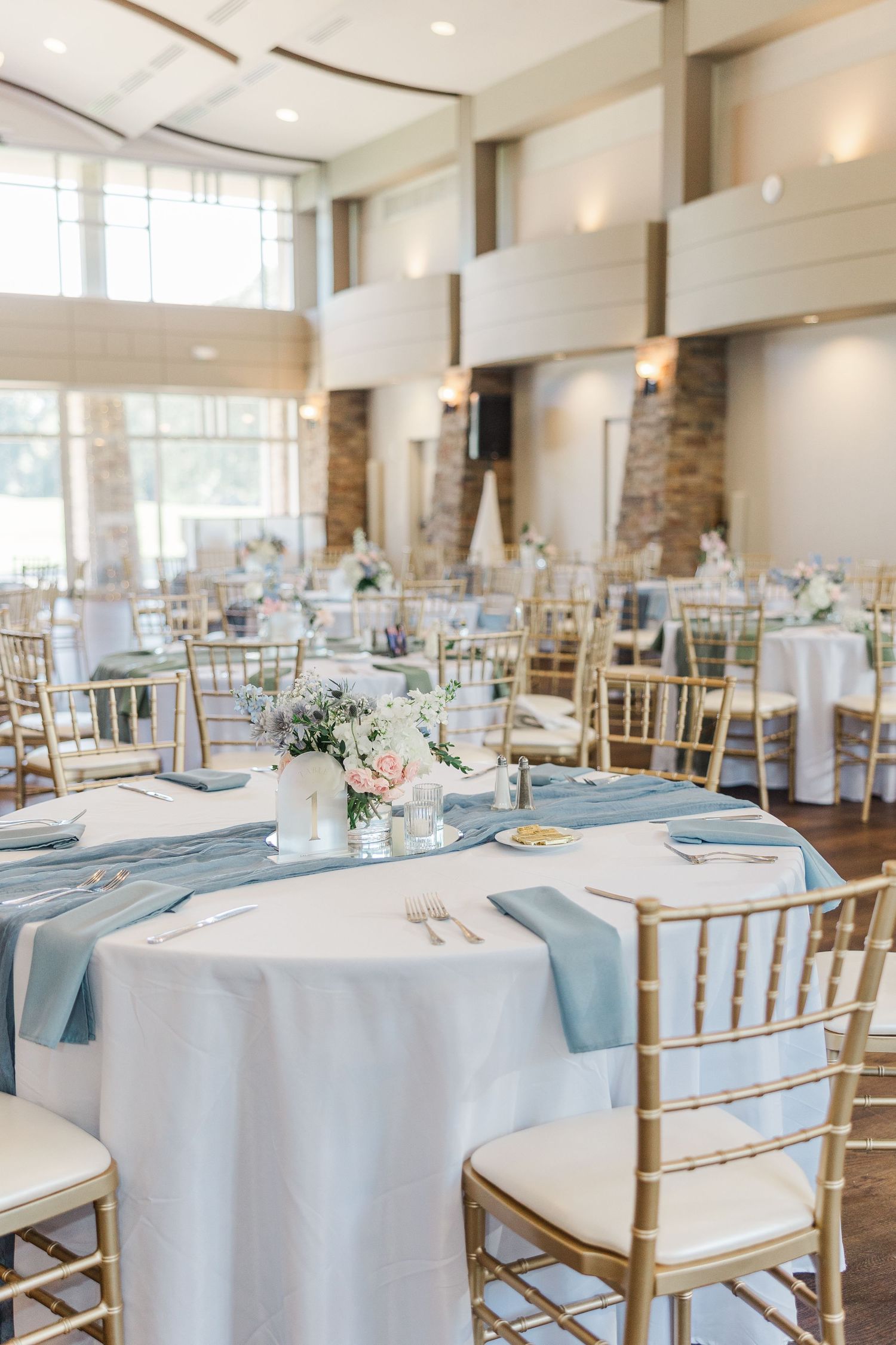 Elegant wedding reception hall with gold Chiavari chairs, light blue table runners, white linens, and floral centerpieces.