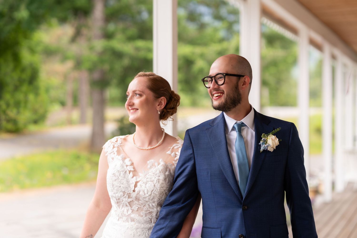 Happy newlyweds smile together outdoors, bride in lace gown and groom in navy suit with boutonniere.