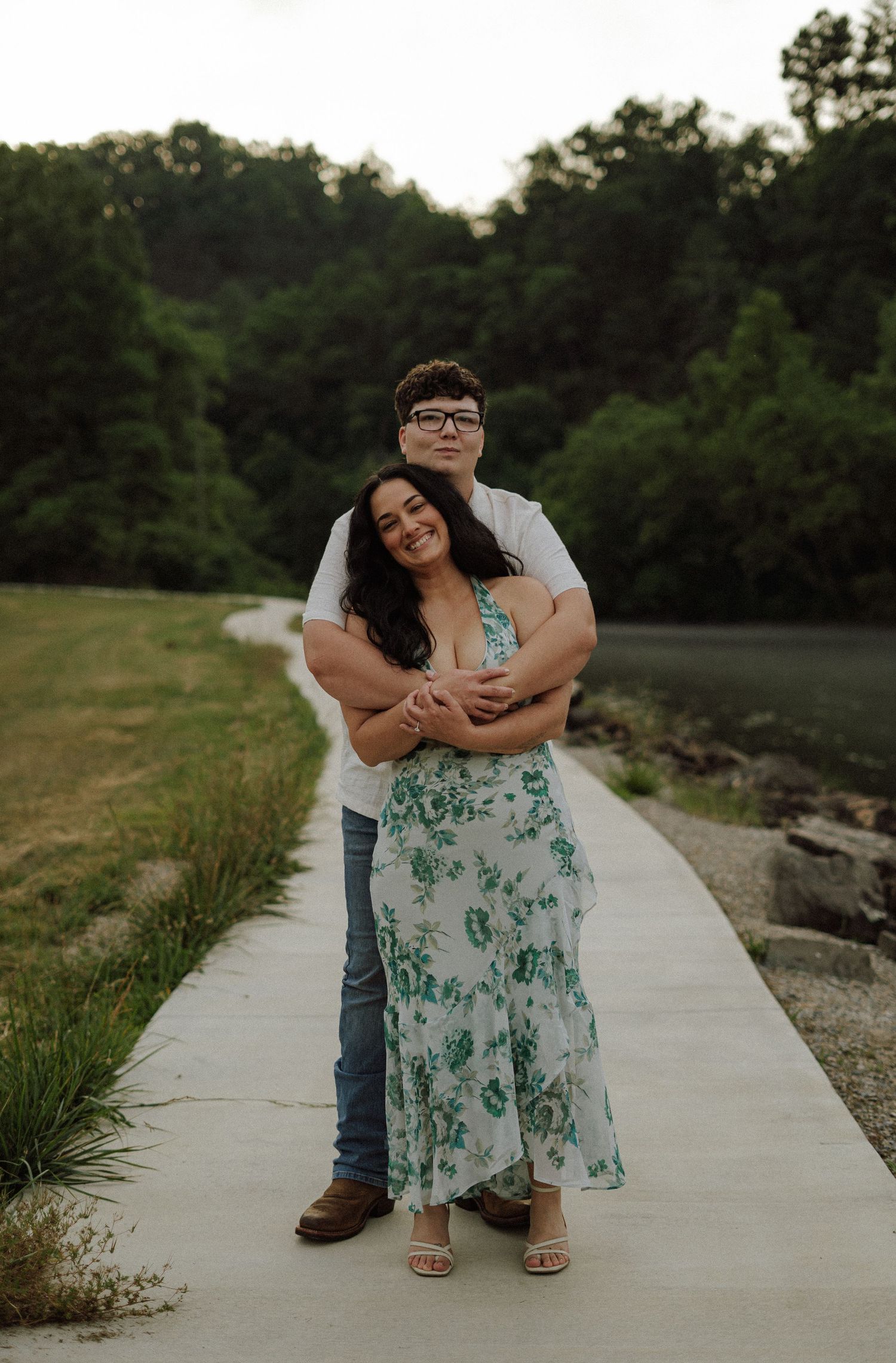Couple embracing on a lakeside path, woman in floral dress and man in white shirt, surrounded by lush green trees.