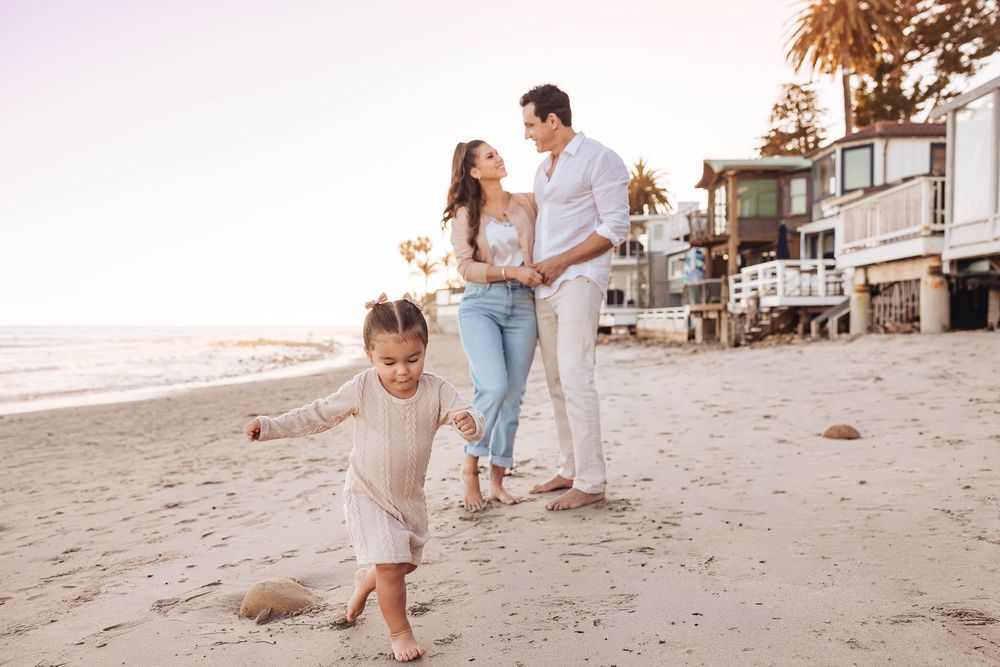 Family enjoying a sunset beach walk, parents holding hands while young child plays in the sand near coastal homes.
