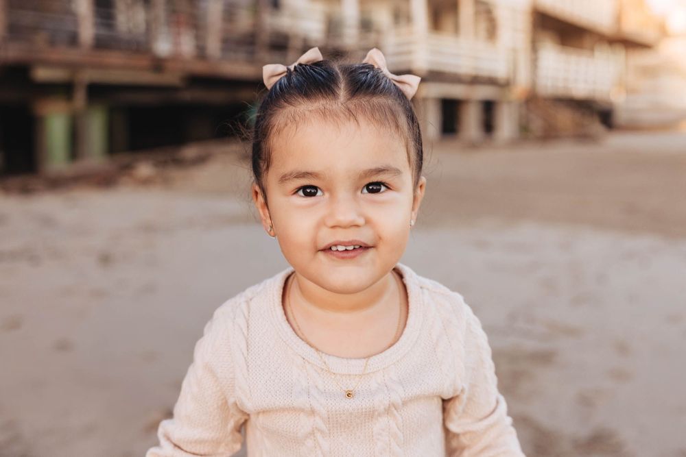 Smiling toddler girl with bow pigtails wearing a cream sweater at a sandy beach location.