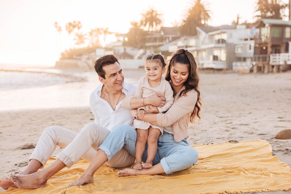 Family of three sitting on yellow blanket on beach at golden hour, parents smiling at toddler.