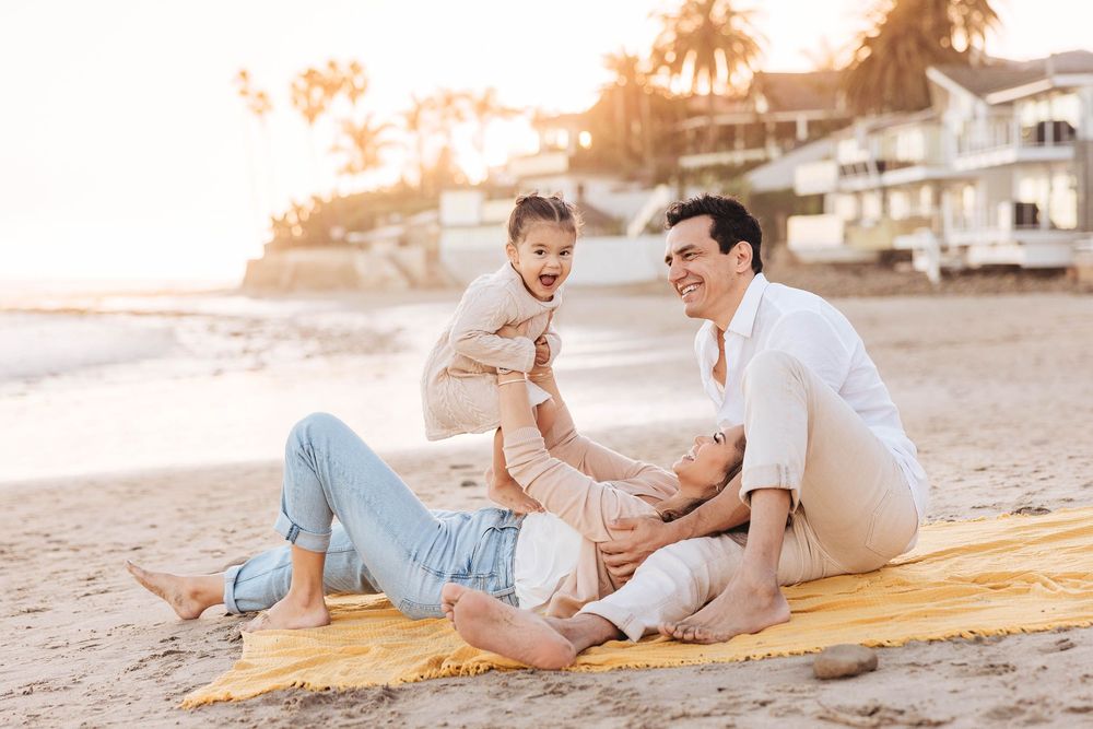 Father playing with laughing toddler on beach blanket at sunset while mother reclines nearby.