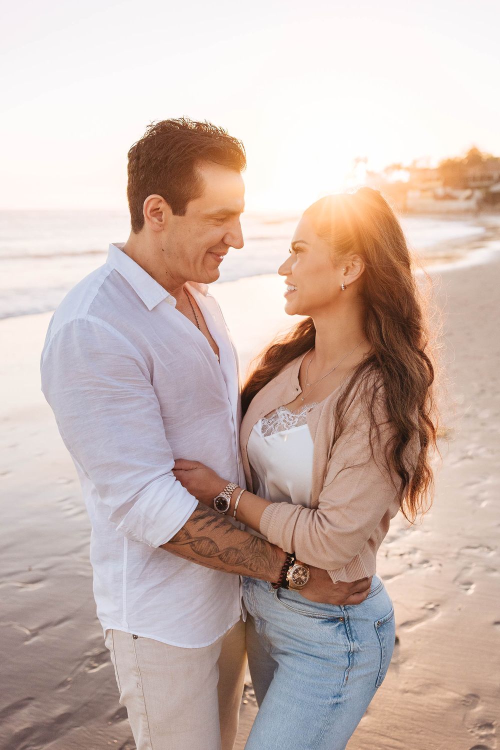 Couple embracing on a sandy beach at golden sunset, woman with long brown hair in jeans, man in white linen shirt.
