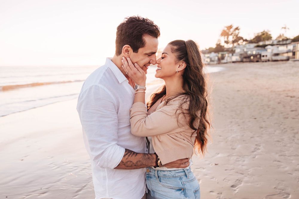 Romantic couple sharing a tender moment on a sunny beach, woman holding man's face while smiling.