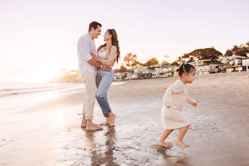 Couple sharing a romantic moment on sunny beach while their toddler splashes in shallow water.
