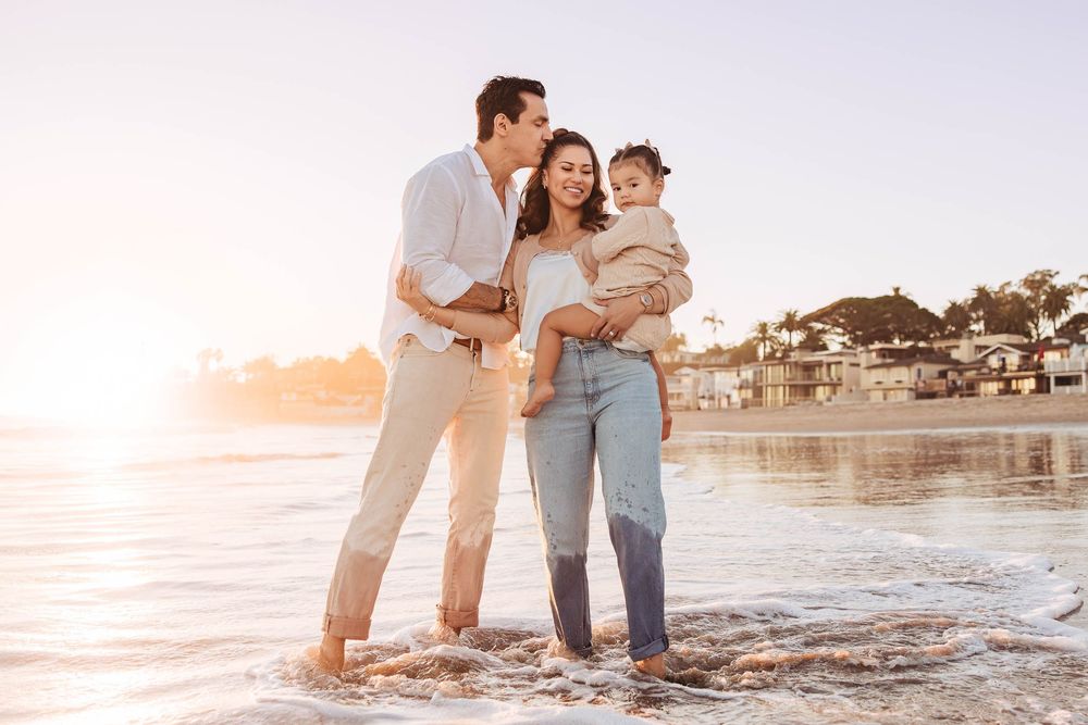 A happy young family with a baby stands on a sunny beach at golden hour, waves lapping at their feet.