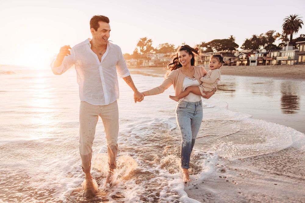 Happy family walking on sunny beach at sunset, parents holding hands with baby, waves splashing at their feet.