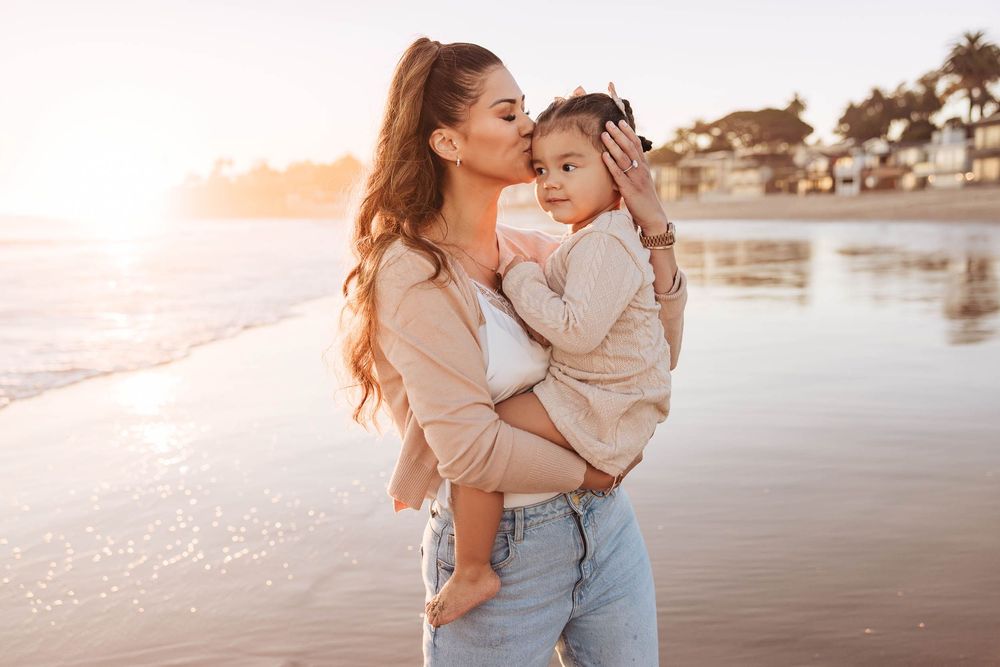 A mother kisses her young daughter on the beach at golden hour, both wearing neutral tones near the ocean shore.