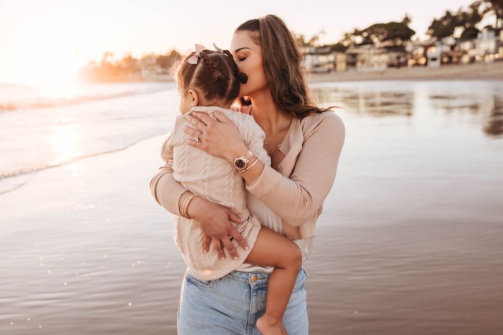 Mother kissing her young daughter on the cheek while holding her on a sunny beach at sunset.