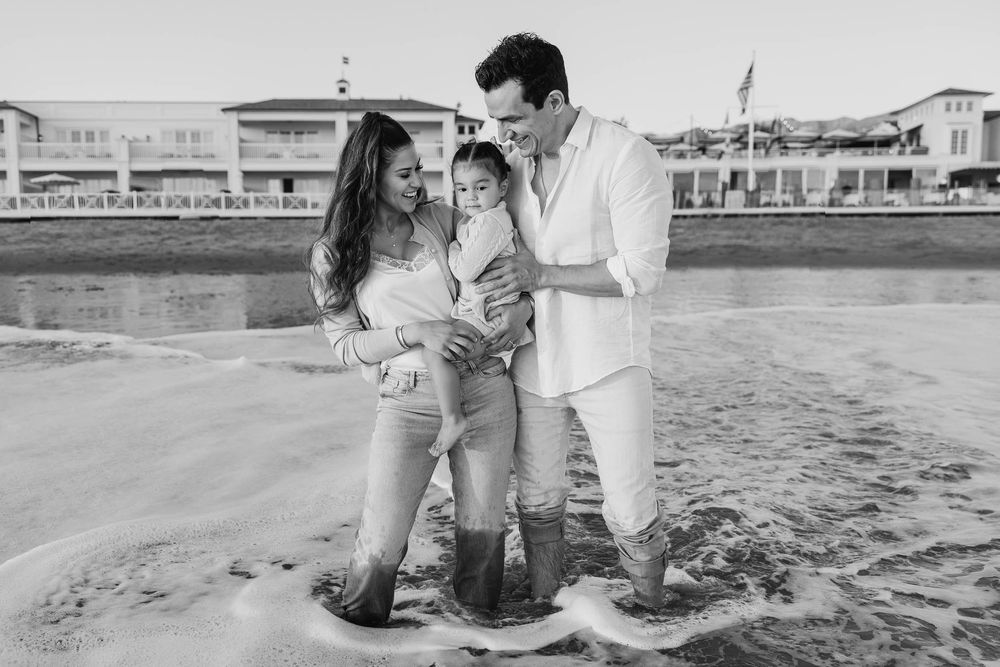 Black and white photo of family of three standing in ocean waves near beach houses at sunset.