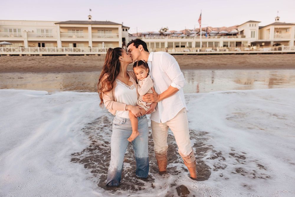 A couple sharing a kiss over their baby while standing in ocean waves on a sandy beach near a resort.