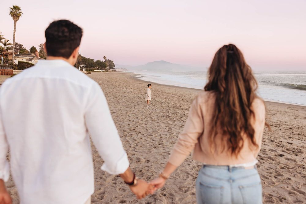 Couple holding hands walking on a beach at sunset with child playing in the distance.