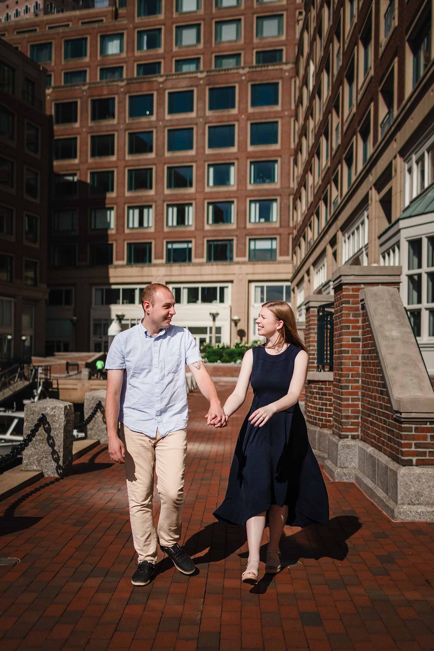 Engaged couple holding hands and walking along the Harbor Walk near Rowes Wharf in Boston