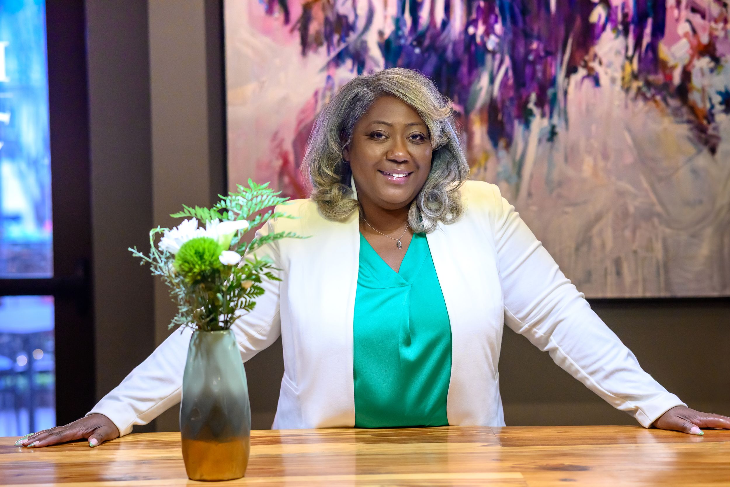 Professional woman in white blazer and green top smiling confidently at wooden table with floral arrangement.