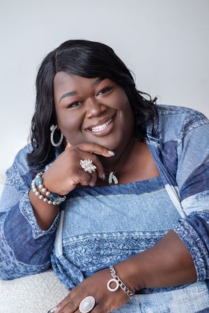 Smiling Black woman in blue denim top wearing silver jewelry, bracelets, and rings, posing confidently against light background.