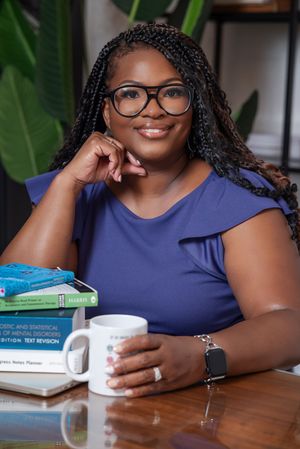 A smiling woman in a blue top sits at a desk with psychology books and a coffee mug, wearing glasses and braids.